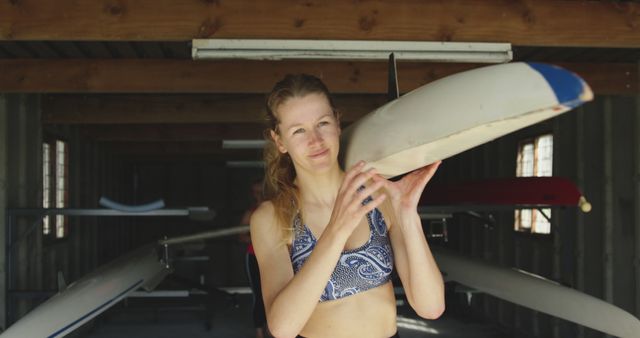 Young woman holding kayak paddle in a boathouse. She seems focused and determined, suggesting she is preparing for a kayaking session. This image can be used for promoting water sports, fitness programs, or healthy lifestyles. It's suitable for marketing sports gear, outdoor adventure tourism, or motivational content.