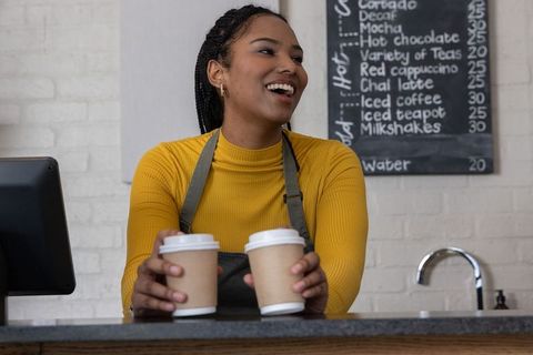 African american barista smiling handing two to-go coffees at cafe counter with chalk menu