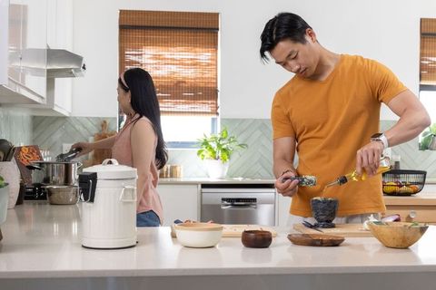 Asian couple cooking together in modern bright kitchen, man pouring oil into mortar bowl