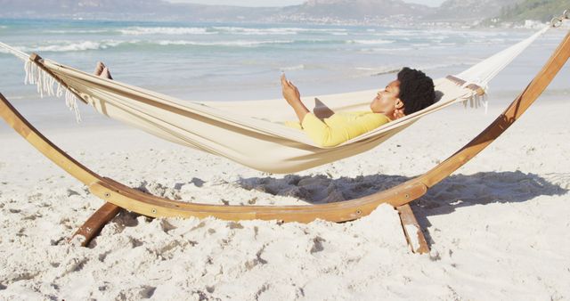 Happy african american woman reading and lying in hammock on sunny beach. healthy and active time beach holiday.