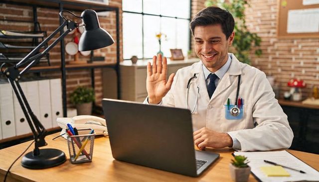 Smiling doctor with stethoscope waving during telemedicine video call in modern clinic office