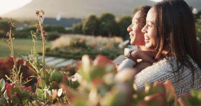 Happy romantic biracial lesbian couple sitting and embracing in garden at sundown, slow motion. Lifestyle, relationship, togetherness, love, relaxation and free time, unaltered.