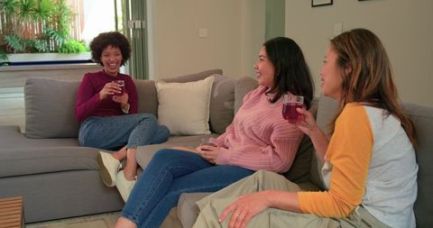 Multiethnic friends laughing and chatting on grey sofa while enjoying drinks at home