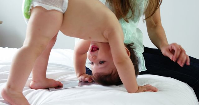 A young Caucasian woman plays with a baby, lifting the child upside down on a bed, with copy space. Their playful interaction captures a moment of joy and bonding between caregiver and child.