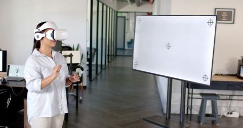 Woman wearing vr headset exploring virtual interface next to blank monitor in office