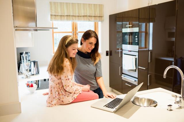 Mother and daughter are using a laptop together in a modern kitchen. The mother is standing while the daughter is sitting on the kitchen counter. Both are smiling and appear to be enjoying their time together. This image can be used for themes related to family bonding, technology use at home, modern lifestyle, and parenting. It is suitable for websites, blogs, and advertisements focusing on family life, home activities, and educational content.