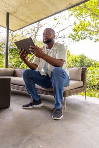 African man sitting on rattan sofa holding tablet on covered patio