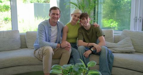 Smiling modern family relaxing on light sectional sofa in sunlit living room with greenery