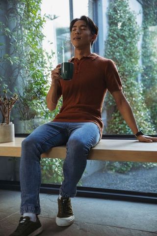 Asian man sitting by window holding mug and savoring coffee aroma in sunlit green space