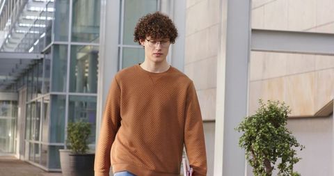 Student walking with notebooks outside campus building wearing glasses and quilted brown sweater
