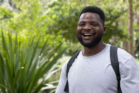 Laughing man with backpack enjoying sunny park stroll, candid portrait, greenery
