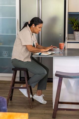 Mature woman working at kitchen island with laptop and earbuds, focused remote work day