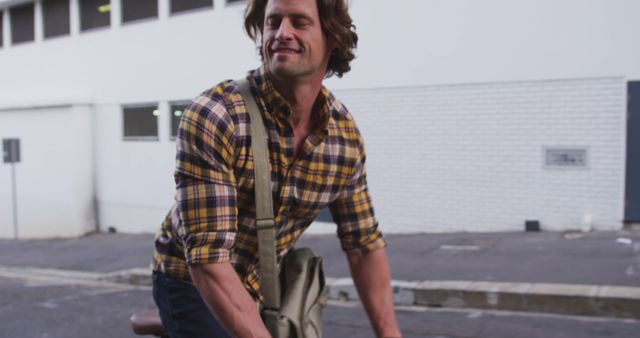 Man riding bicycle on urban street, wearing plaid shirt and shoulder bag, smiling casually. Perfect for themes on urban lifestyle, transportation, travel, leisurely commutes, or promoting eco-friendly commuting options.