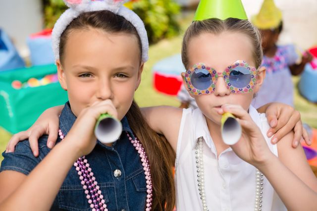 Two young girls are celebrating a birthday party outdoors. They are blowing party horns and wearing festive accessories like party hats and necklaces. This image can be used for themes related to children's parties, celebrations, friendship, and outdoor fun.