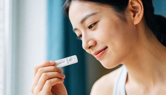 Young woman reading pregnancy test by window, smiling in quiet hopeful home moment