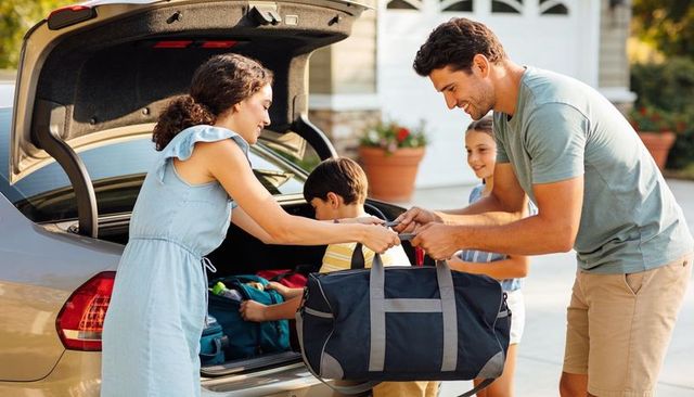 Family packing car for weekend getaway, parents loading duffel into trunk while kids help