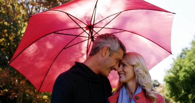 A middle-aged Caucasian couple shares a tender moment under a vibrant red umbrella, with copy space. Their affectionate gaze and smiles suggest a romantic connection as they enjoy a sunny day outdoors.
