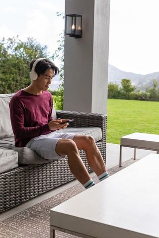 Asian man sitting on rattan patio sofa wearing headphones and using smartphone in outdoor lounge