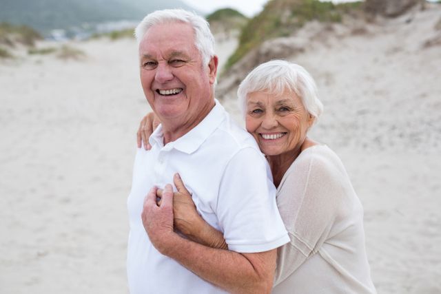 Senior couple embracing on sandy beach, both smiling joyfully. Ideal for promoting retirement living, senior travel, lifestyle, and health or relationship content.