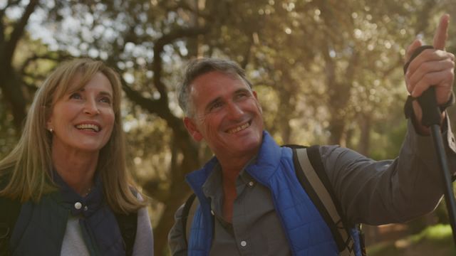 This image captures a joyful senior couple enjoying a hike in a lush forest, both smiling and interacting with nature. Ideal for use in content promoting active lifestyles, senior adventures, outdoor activities, personal well-being, or tourism targeting older adults. It conveys themes of happiness, vitality, and appreciation of the outdoors.