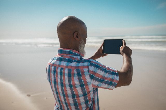 Senior African American man standing on beach taking picture with tablet. Ideal for themes related to summer vacations, leisure activities, technology use among seniors, and beach holidays.