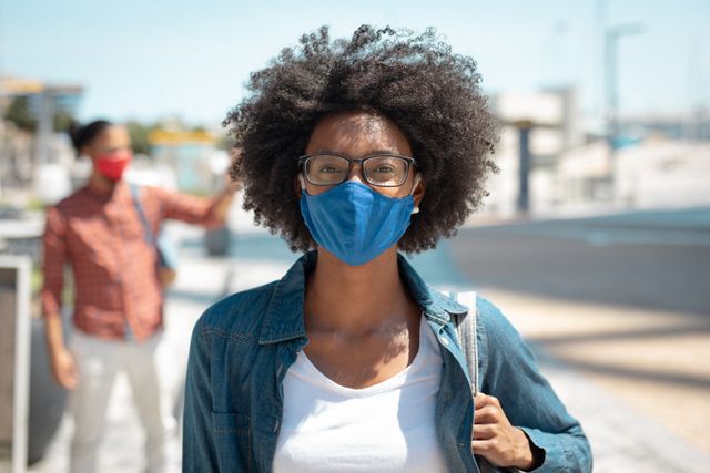 African American woman wearing a face mask and glasses, walking in a sunny urban street. Ideal for illustrating themes related to COVID-19 safety, public health, urban lifestyle, and pandemic precautions. Suitable for use in articles, advertisements, and social media posts promoting health and safety measures.