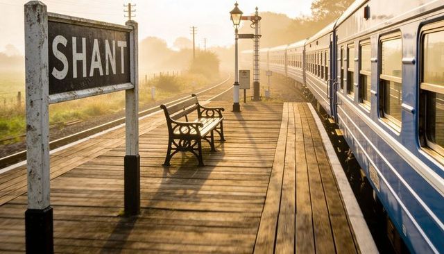 Gliding blue train curving past rustic wooden platform with bench and vintage station sign
