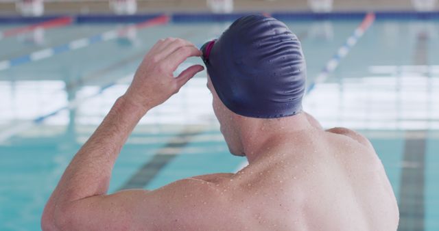Athlete adjusts swimming goggles and swim cap before starting workout at indoor swimming pool. Ideal for use in fitness, sports, swimming tutorials, health and wellness projects. Captures dedication and preparation in athletic training.