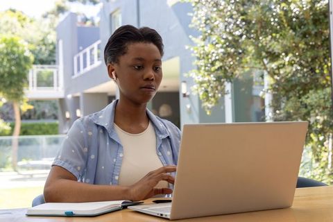 Focused black teen working on laptop outdoors at modern home patio with earbuds and notebook
