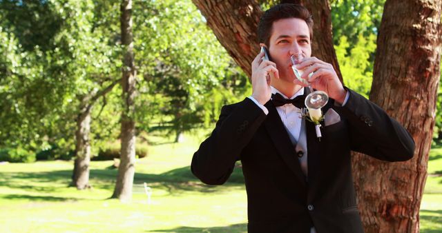 Groom wearing black suit and bow tie talks on phone and drinks water outdoors surrounded by trees and green grass. Suitable for use in wedding blogs, articles about wedding preparation and communication, or advertisements for wedding services.