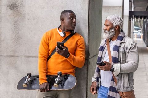 African american young man and senior talking on sidewalk with skateboard and smartphones