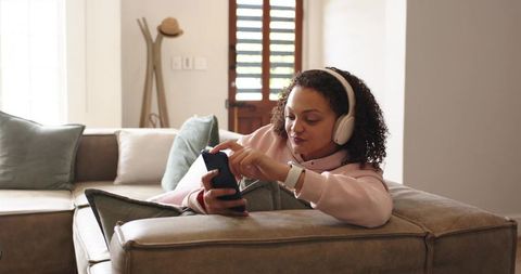African american woman reclining on sectional sofa using smartphone and wireless headphones