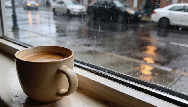 Steaming coffee cup on rain-streaked city windowsill with crema and street reflections