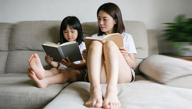 Mother and daughter reading on gray sofa barefoot, cozy home bonding and relaxation