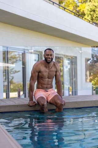 African american man sitting on pool deck dipping feet in water in peach swim trunks