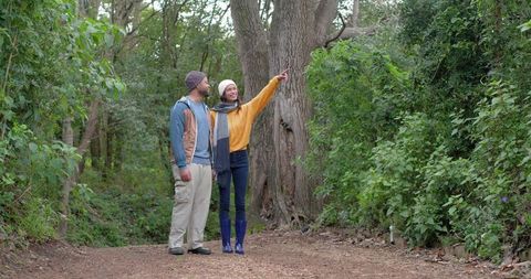 Diverse couple exploring forest trail pointing at tree, wearing knit hats and rain boots