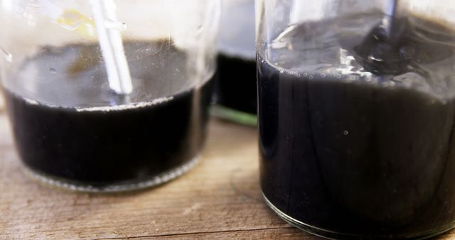 This image effectively captures a detailed close-up of a thick, black liquid being poured into glass jars on a wooden surface. Perfect for uses in articles, marketing materials, or advertisements related to food, cooking, crafting, laboratory work, or any context that involves thick fluids, such as syrup, oil, or other substances. The image creates a mysterious and intriguing atmosphere with its dark tones and the rich texture of the liquid.