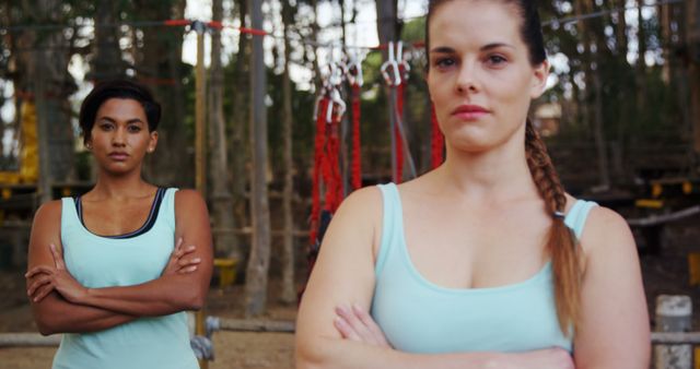 Two female athletes posing confidently with arms crossed at an outdoor gym. Background shows gym equipment and a natural forest setting, indicating a focus on outdoor fitness and wellness. Perfect for use in fitness-related content, promotional material for gyms or athletic programs, and articles promoting women's empowerment in sports.