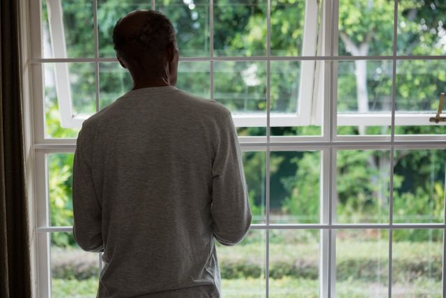 This image depicts a senior man standing by a window, looking outside. It can be used in articles or advertisements related to retirement, elderly care, solitude, contemplation, and peaceful living. The natural light and greenery outside add a serene and calm atmosphere, making it suitable for wellness and mental health topics.