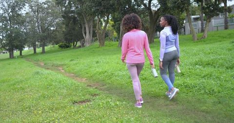 Two women walking on grassy park trail in activewear carrying water bottles for wellness