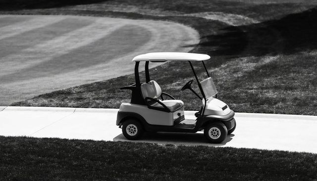 Empty golf cart parked on cart path beside manicured fairway with mowing stripes in monochrome