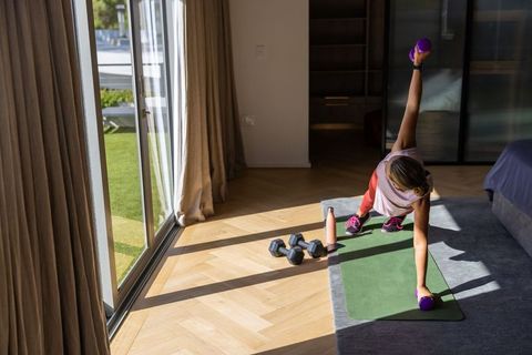 Woman performing side plank with dumbbells on yoga mat in bright bedroom workout space