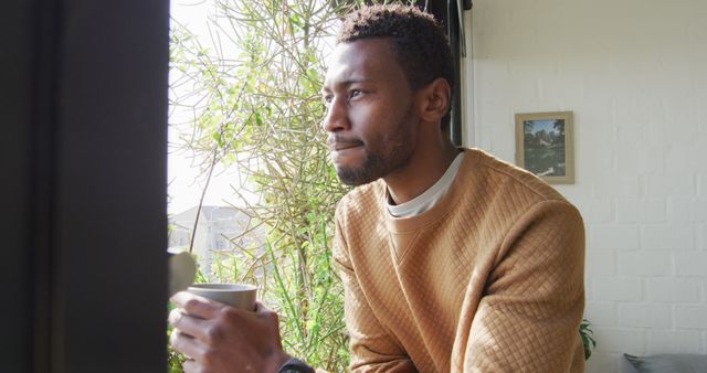 Man wearing light brown sweater holding coffee cup looking out of window. Room features white brick wall and potted plant. Great for themes of relaxation, morning routines, work breaks, or reflective moments. Ideal for use in lifestyle blogs, mental health awareness, and advertisements for home products.