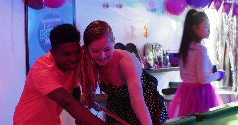 Young diverse friends playing pool at lively birthday party with neon lighting