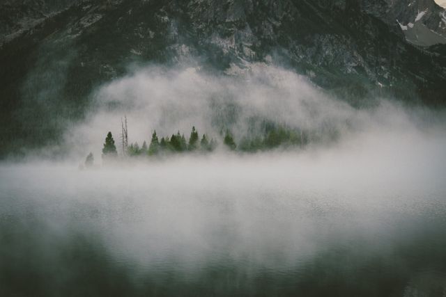 Ethereal scene showing small island covered in trees, surrounded by mist at mountain lake. Perfect for themes of tranquility, nature, serenity, and wilderness. Excellent for travel blogs, meditation materials, serene landscape features, and nature photography collections.