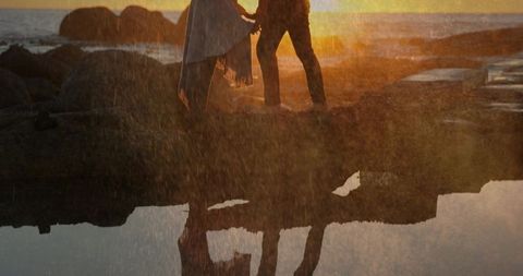Silhouetted couple standing on rocky shore at sunset with tidal pool reflection