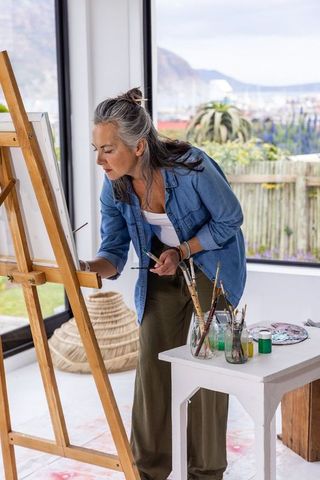 Mature woman painting at wooden easel in sunlit studio with ocean view, holding brush