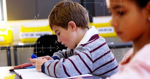 Young focused boy concentrating while writing math notes in classroom with classmate nearby