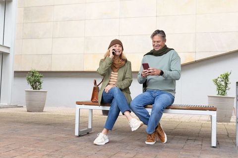 Mature couple sitting on urban bench, smiling and using smartphones, autumn casual style