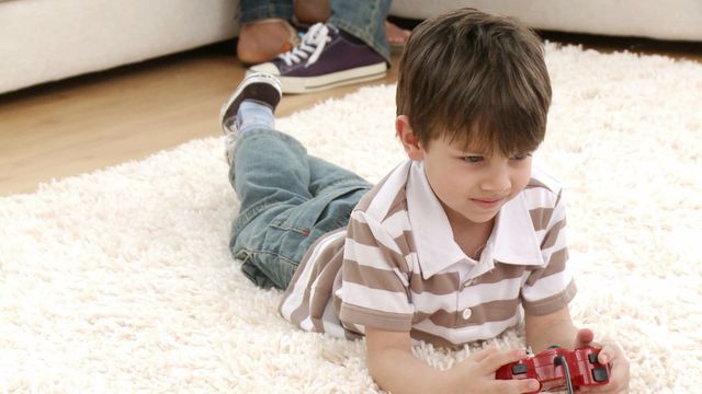 Young boy lying on a carpet, playing a video game with family present in the living room. Great for themes of family bonding, children's activities, indoor leisure, and cozy home environments.